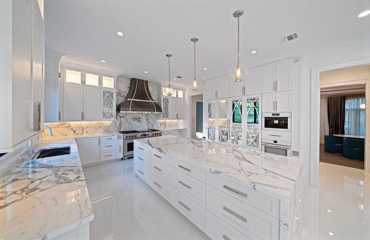 White marble kitchen with a large island, range hood, and glass-front cabinets